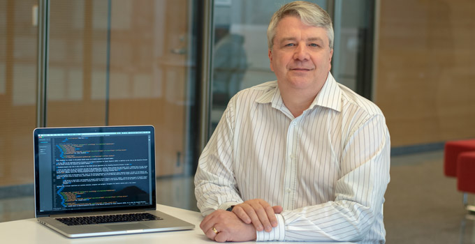 Dr. John Bartlett poses for a photo at a table next to a laptop computer displaying lines of code. 